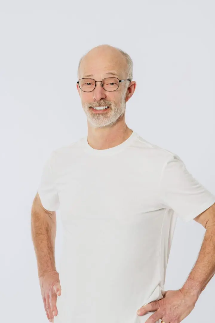 A man with glasses and white shirt standing in front of a wall.